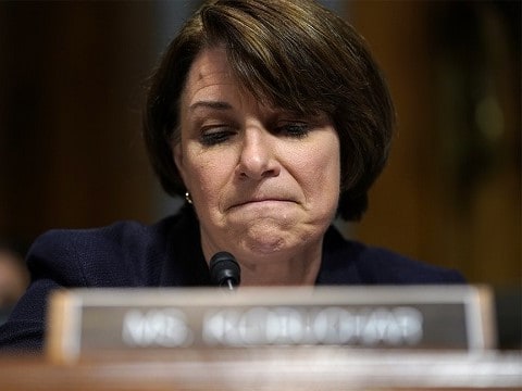 minnesota senator amy klobuchar sitting behind a name placard frowning