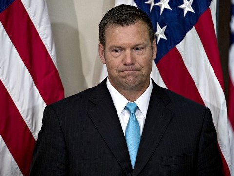 kris kobach frowning while standing in front of an american flag